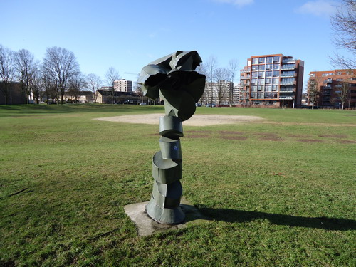 De omgehakte boom Alexanderplantsoen
Een monument voor een omgehakte boom. Je moet toch een groot hart hebben, wil je dat maken. Dat had Nico Betjes. Ondanks zijn robuuste uiterlijk en tegenover onbekenden soms wat norse houding, was het een uiterst aimabel mens. Een mens ook met een hart voor natuur. Zijn monument voor een omgehakte boom lijkt op het eerste gezicht een volstrekt abstract object. Dit monument is in 1975 is gemaakt en staat aan de Laan der Nederlanden schuin tegenover de Vredevorstkerk. Aan de titel van dit monument  zie je al snel dat het een in blokken gehakte stam voorstelt die Betjes weer rechtop heeft gezet. In een spiraalvorm, die wellicht ‘beweging’ of groei voorstelt. Bovenop staat een nieuwe kruin. Mooi is ook dat de boom opnieuw lijkt om te vallen. Het beeld spreekt veel Beverwijkers enorm aan. Tijdens de renovatie van de wijken Oosterwijk/ Meerestein is het lange tijd van zijn plek geweest. In die periode is het beeld ook gerestaureerd. Toen er op een gegeven moment sprake was van een van een verhuizing van her beeld naar een andere plek in Beverwijk, kwam het wijkcomité in opstand, zo’n beeld wil je niet meer kwijt. 

Bron: Blik op Beeld, Tekstbureau Zonneveld & Thöne i.o.v. gemeente Beverwijk, 2006.
Foto en bewerking: Co Backer
Keywords: bwijk Openbare Kunstwerken