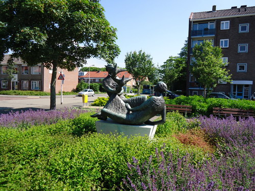 Mens-stad-leven
Een nieuwe tijd, een nieuw beeld. Deze beeldengroep straalt optimisme uit en geloof in de toekomst. Het werd gemaakt in een tijd waarin alles nieuw leek te worden. Beverwijk had nog ruimte in overvloed en de welvaart steeg met sprongen. De stad vernieuwde zich voortdurend. Nieuwe woonwijken, scholen bedrijven, en een nieuw stationsgebouw. Die vernieuwingen vonden hun bekroning in de plaatsing van een eigentijds beeldhouwwerk. Dat was het realistische ‘Mens-stad-leven’. De recht opzittende figuur is ‘de cultuur’ die Beverwijk, de zittende figuur, uit haar slaap wekt. De opvliegende duiven verbeelden op hun beurt de vrije uitbreiding en ontwikkeling van de stad. De kunstenares, Annemarie Leenders-Kusters, heeft meerdere beelden voor Beverwijk gemaakt, waaronder de Tuinder ofwel De Schoffelaar. Op één en dezelfde dag in 1960 werd de beeldengroep onthuld en het nieuwe stationsgebouw en het nieuwe stationsplein feestelijk geopend. Doordat het Stationsgebied opnieuw wordt ontwikkeld is deze beeldengroep tijdelijk verplaatst naar de hoek Huiderlaan / Wilgenhoflaan.  

Bron: Blik op Beeld, Tekstbureau Zonneveld & Thöne i.o.v. gemeente Beverwijk, 2006.
Foto en bewerking: Co Backer
Keywords: bwijk Openbare Kunstwerken