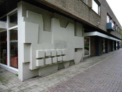 Openbare Bibliotheek Kerkstraat
Vanaf 1950 ging het hard met het plaatsen van kunstwerken in de openbare ruimte in Nederland. Niet omdat er bij elke willekeurig gemeentebestuur plotsklaps het besef doordrong van het belang van kunst. Nee, het was de 1-procentsregeling van de overheid die deze opvallende toenamen van kunstwerken verklaart. Volgens deze stimuleringsmaatregel moest 1% van de bouwsom van elk nieuwbouwproject besteed worden aan kunst. Gemeenten werden er overigens niet aangehouden om mee te doen.  Beverwijk paste deze regeling vooral toe in de jaren zestig en zeventig van de vorige eeuw. Na 1980 raakte deze regeling in Beverwijk in onbruik. Na voltooiing in 1970 van het bibliotheekgebouw aan het Kerkplein koos het Beverwijks gemeentebestuur voor Piet Schoenmakers uit Roermond om een kunstwerk te maken. Het werd een zogenoemd bas-reliëf  aan de gevel. Het heeft geen titel mee gekregen, omdat men er in mag zien wat men wil. Het verbeeldt niets, het is uitsluitend de vorm die moet bekoren. En de structuur, want wie goed kijkt ziet dat iedere laag een andere oppervlaktestructuur heeft. Een ijverige schilder had het bas-reliëf  ooit kleuren gegeven. Nu is het weer in oorspronkelijke staat en dat is naturel. Meer van Piet Schoenmakers in Beverwijk: Weerhaan aan de Plesmanweg en de Levenscyclus die boven de ingang van het oude Stadkantoor hing.       

Bron: Blik op Beeld, Tekstbureau Zonneveld & Thöne i.o.v. gemeente Beverwijk, 2006.
Foto en bewerking: Co Backer
Keywords: bwijk Openbare Kunstwerken