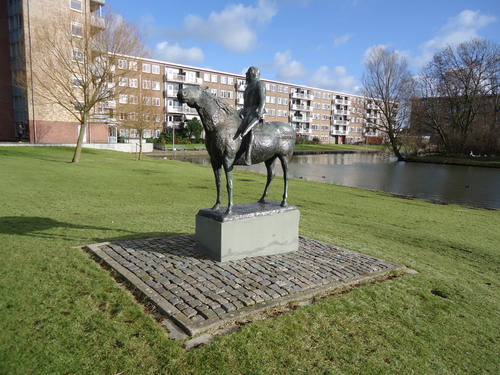 Paard met ruiter Laan der Nederlanden
Ooit geweten dat op het eiland in de vijver resten van een middeleeuws Kasteel liggen?  Toen kunstenaar Eric Claus hiervan hoorde liet hij zich, voor de opdracht om een beeld op deze plek te vervaardigen, door deze kennis inspireren. Hij schiep ‘Paard met ruiter’ dat bijna als vanzelf meeleven oproept. De vermoeidheid straalt van het paard en zijn metgezel af. De strijd is gestreden. Op naar huis. Het is dat het beeld op zo’n hoge sokkel staat, maar anders zou je er toch graag even aan komen. De commissie die destijds Claus uitkoos om een beeld te vervaardigen voor het nieuwe plan ‘Oosterwijk’  had een vooruitziende blik. Claus, jong nog, had net zijn eindexamen aan de Academie voor Beeldende Kunsten te Amsterdam afgerond. Dit beeld was zijn deerste grote opdracht. Daarna zijn er van deze veelzijdige en productieve kunstenaar vele beelden in Nederland en ook in Amerika geplaatst. Het paard met ruiter maakte deel uit van een serie kunstopdrachten gegeven door wijlen burgemeester  Bruinsma, waarbij een viertal kunstenaars een beeldhouwwerk moest maken voor het nieuwe plan ‘Oosterwijk’. Dit beeld is onthuld in 1964 en staat aan de Laan der Nederlanden, nabij de vijver. 

Bron: Blik op Beeld, Tekstbureau Zonneveld & Thöne i.o.v. gemeente Beverwijk, 2006.
Foto en bewerking: Co Backer
Keywords: bwijk Openbare Kunstwerken