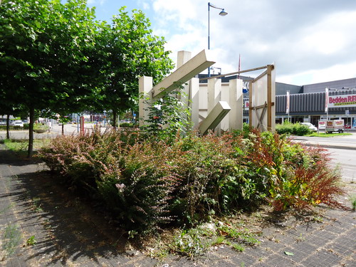 Staal op een betonnen sokkel Meubelb.
Ooit bewogen de afzonderlijke horizontale delen van dit beeld, dat staat op de Parallelweg voor het toenmalige wooncentrum Stoutenbeek, op elk zuchtje wind dat er langs streek. Eigenaar Stoutenbeek vond dat echter te gevaarlijk voor passerende kinderen. Zonder slag of stoot liet hij de onderdelen vastlassen. Ondanks dat statische blijft het een opvallend beeld, en dat was precies de bedoeling van de maker Dick Box. De plaats van de ingang van de zaak was de vorm van de gevelarchitectuur niet direct duidelijk. Box koos ervoor om het markante kunstwerk pal voor de ingang te zetten. De beweeglijkheid van de afzonderlijke delen contrasteerde mooi met het strakke pand. Het beeld is opgebouwd uit negen vierkante buizen en heeft telkens een veranderde lijnenspel. Volgens de maker zit de essentie van het beeld in het stabiele versus het instabiele. Helaas, de eigenaar besliste anders. Het in 1978 geplaatste beeld kijkt nu anno 2013 nog net boven het onkruid uit en is aan de straatkant “afgeschermd” met twee borden die het pand aanprijzen aan een mogelijke koper/huurder.    

Bron: Blik op Beeld, Tekstbureau Zonneveld & Thöne i.o.v. gemeente Beverwijk, 2006.
Foto en bewerking: Co Backer
Keywords: bwijk Openbare Kunstwerken